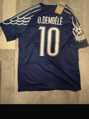 PSG fotbollströja O. Dembélé #10 - Säljer en mörkblå PSG fotbollströja med O. Dembélé och nummer 10 på ryggen. Tröjan har vita detaljer, vingmönster på axlarna, Qatar Airways-logga på bröstet och Champions League Foundation-märke på ärmen. Materialet är lätt polyester och har Jordan-loggan på framsidan.
