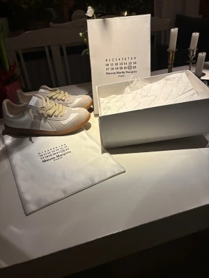  Margiela gats - Maison Margiela sneakers i beige och vit med klassisk gummisula. Skorna har rund tå, snörning och detaljer i både skinn och mocka. Medföljer originalkartong, dustbag och StockX-verifiering. Perfekt för dig som gillar stilrena och lyxiga sneakers. Använda ett fåtal gånger men är i fel storlek så drf jag säljer, pris kan diskuteras vid snabb affär :)