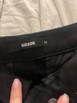 Svarta wide kostymbyxor från Bikbok - Snygga svarta kostymbyxor från Bikbok i storlek 34. Byxorna har en wide passform med raka, vida ben och klassiska fickor fram. Perfekt för dig som gillar stilrena och trendiga plagg. Materialet känns mjukt och har en lätt glans. Modellen Vera