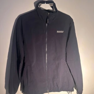 Woolrich windbreaker  - Mörkblå vindjacka från Woolrich med hög krage och hel dragkedja framtill. Jackan har två sidofickor med dragkedja och diskret logga på bröstet. Materialet är lätt och känns som polyester, perfekt för blåsiga dagar. Klassisk och stilren design med normal passform.