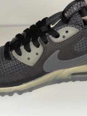 Nike Air Max Terrascape grå sneakers - Nike Air Max Terrascape sneakers i mörkgrå och ljusgrå toner med rutmönstrad textil och detaljer i syntet. Swoosh-logga på sidan, svart snörning och transparent sula med Air Max-dämpning. Insidan har limegrön innersula med Terrascape-tryck.