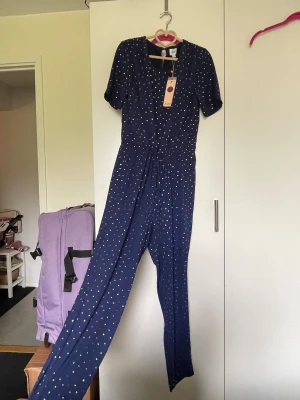 Mörkblå prickig jumpsuit från Saint Tropez - Snygg mörkblå jumpsuit från Saint Tropez med vita prickar, korta ärmar och rund hals. Byxdelen är rak och har normal passform, med knappar i ryggen och ett band i midjan för extra stil. Perfekt för dig som gillar trendiga och bekväma plagg.Helt ny med pris lapp. Inköpt pris 699kr