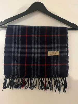 Burberry halsduk - Stilren halsduk från Burberry i mörkblå, grå och röd färg. Halsduken är tillverkad av 100% lammull och har klassiska fransar längst ner. Perfekt just nu till vintern och kallare dagar. Skriv vid minsta fundering!