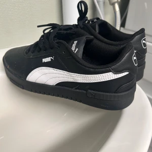 Svarta och vita Puma sneakers strl 38.5 - Säljer ett par klassiska Puma sneakers i svart och vitt med snygg logga på sidan och hälen. Skorna har snörning, platt sula och är gjorda i syntetmaterial med läderlook. Jag har använt skorna max tre gånger. De är för platta för mina fötter där av säljer jag de. 