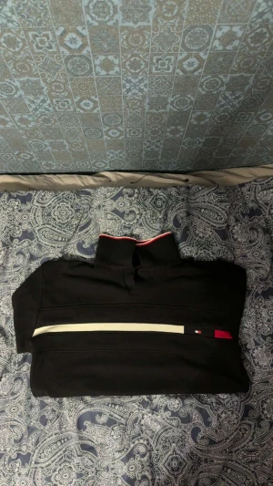 Svart half zip tröja Tommy Hilfiger - Svart långärmad half zip tröja från Tommy Hilfiger med vit och röd rand över bröstet och liten logga. Hög krage med röd och vit kant. Perfekt för dig som gillar stilren och sportig look. Den är slimfit så den passar både M och S