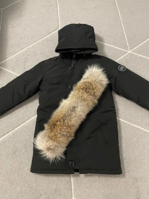 Chateau Canada Goose  - Svart dunjacka från Canada Goose med huva och avtagbar pälsdetalj i beige och bruna toner. Jackan har stora knappar, dragkedja och två rejäla fickor framtill. Insidan är quiltad och jackan har en patch med logga på ärmen.