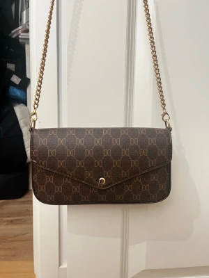 Brun axelväska från Don Donna - Axelväska från Don Donna (Glitter) med kedja i guld (som är rostig, se bilderna) och monogrammönster. Perfekt storlek för mobil, plånbok och småsaker.