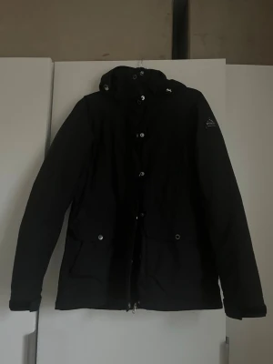 Svart jacka från McKinley, strl 38 - Svart jacka från McKinley med huva och dragkedja framtill. Jackan har stora fickor med knappar och är helsvart med en stilren look. Materialet är polyester. Jackan har en innerjacka som enkelt kan tas i och ur beroende på hur tjock jacka som önskas. Mycket fint skick!