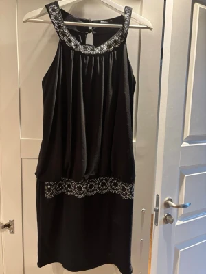 Svart kort klänning Bodyflirt 38 - Snygg svart klänning från Bodyflirt i storlek 38. Klänningen har ärmlös design med glittriga detaljer vid halsringningen och midjan. Den har en avslappnad passform och är perfekt för dig som gillar att sticka ut med lite extra bling.