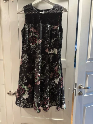 Blommig svart klänning från Rainbow - Snygg svart klänning från Rainbow med blommigt mönster i rosa, grönt och vitt. Ovanpartiet har dekorativ spets och klänningen är ärmlös med rund hals. Kjolen är lätt utsvängd och klänningen är i kort modell, perfekt för dig som gillar detaljer och blommigt.