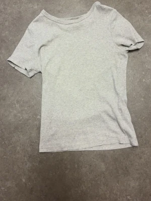 Ljusgrå ribbad t-shirt XS - Säljer en ljusgrå ribbad t-shirt i storlek XS. T-shirten har korta ärmar och en klassisk rund halsringning. Materialet är mjukt och stretchigt, perfekt för en chill och enkel stil. Passar dig som gillar basic och cleana plagg.