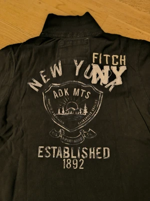 Abercrombie & Fitch henley  pike - Svart henley från Abercrombie & Fitch med tryck och broderade detaljer på ryggen och ärmen. Röd logga på bröstet och vit älg på vänster ärm. Piken har korta ärmar, krage och knappar framtill. Perfekt för dig som gillar street och amerikansk college-vibe.