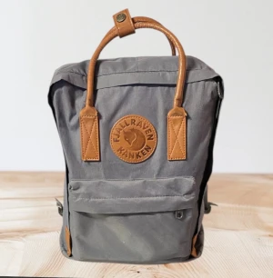 Grå Fjällräven Kånken ryggsäck - Fjällräven Kånken Classic ryggsäck i grått med bruna ej läderdetaljer. Väskan har en framficka och justerbara axelremmar. Observera att bilden är mer detaljerad. • höjd 38cm • bredd 27cm • djup 14cm