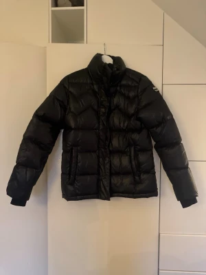 Svart pufferjacka från Mountain Works - Säljer en svart pufferjacka från Mountain Works med hög krage och snygga quiltade detaljer. Jackan har två fickor framtill och logotyp på ärmen. Perfekt för kalla dagar.