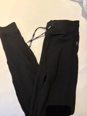 Svarta ribbade träningstights från STRONGER - Svarta träningstights från STRONGER med ribbad struktur och bred resår i midjan. Byxorna har snörning framtill med logotyp på snörena och en avslappnad passform. Perfekta för chill eller träning, mjukt material och snygg look.