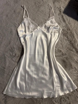 Vitt satin nattlinne med spetsdetaljer - Romantiskt nattlinne i satin med tunna axelband och v-ringad hals. Överdelen har fina spetsdetaljer med blommönster som ger en romantisk vibe. Klänningen har en mjuk, glansig yta och sitter lätt och luftigt på kroppen. Aldrig använd