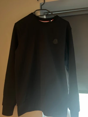 Svart sweatshirt från Moncler - Svart sweatshirt från Moncler i storlek S. Tröjan har rund hals, lång ärm och ett diskret Moncler-märke på bröstet. Klassisk och stilren design med ribbade muddar och insida av kragen med röd, vit och svart detalj. Perfekt för dig som gillar exklusiva streetwear-vibbar. Det går självklart att diskutera pris