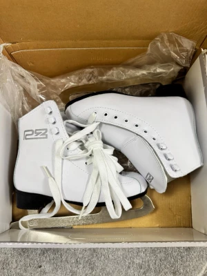 Vita skridskor från PZ med snörning - Snygga vita skridskor från PZ med klassisk snörning och robust svart sula. Skorna har högt skaft och är tillverkade i syntetmaterial med metallblad undertill. Perfekt för dig som vill glida fram på isen med stil. Logga på sidan och detaljerade öglor för snörningen.