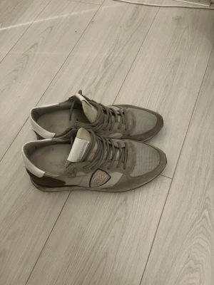 Philippe Model grå sneakers - Säljer ett par snygga Philippe Model sneakers i grå och beige toner med vit häl. Skorna har snörning, rund tå och platt sula. Materialmix av mocka, skinn och mesh. Märkets logga på sidan och bak på hälen. Perfekta för dig som gillar stilrena och exklusiva sneakers.