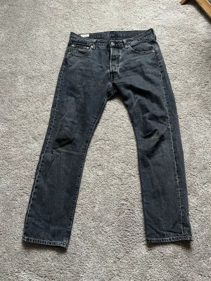 Levi's 501 svarta jeans 34/32 straight fit - Svarta Levi's 501 jeans med klassisk rak passform och fem fickor. Jeansen har knappgylf och är tillverkade i slitstarkt bomullsmaterial. Perfekt för dig som gillar tidlös stil och vill ha ett par jeans som funkar till allt. Märke syns på bakfickan och läderpatch i midjan.