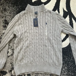 Helt ny Ralph lauren 1/4 zip - Kabelstickad grå tröja från Polo Ralph Lauren med halv dragkedja och hög krage. Klassisk broderad logga på bröstet i mörkblått. Perfekt för lager-på-lager och chill höst- eller vinterstil. Skön passform och ribbade muddar för extra komfort. Obs! Juniorstorlek, passar ålder 14-16 år. 