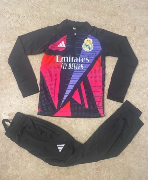 Real Madrid träningsset från Adidas - Säljer ett Real Madrid träningsset från Adidas med långärmad tröja och svarta byxor. Tröjan har coolt grafiskt mönster i rött, lila och svart, samt klubbmärke och Adidas-logga. Byxorna är helsvarta med vit Adidas-detalj. Perfekt för fotbollsträning.