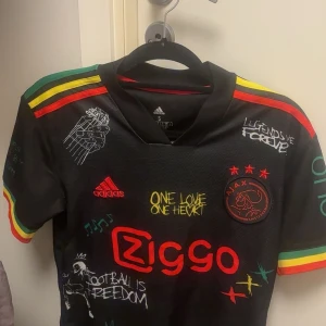 Ajax x Bob Marley Adidas fotbollströja S - Unik svart Ajax fotbollströja från Adidas med Bob Marley-tema. Färgglada detaljer i rött, gult och grönt på axlar och ärmslut, tryck med texter och motiv, samt Ajax-logga och Ziggo sponsor. Tillverkad i lätt och ventilerande material.