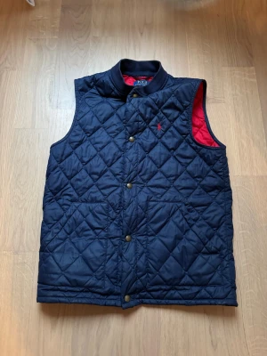 Ralph Lauren Väst - mörkblå - Quiltad mörkblå dunväst från Polo Ralph Lauren med röd insida och tryckknappar framtill. Västen har två stora fickor och ett diskret broderat rött Polo-logo på bröstet. Perfekt för lager-på-lager under höst och vinter.