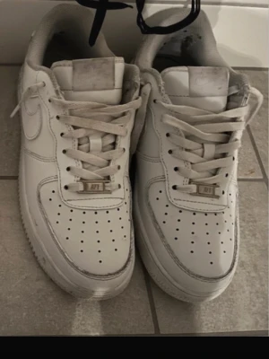Nike Air Force 1 vita sneakers - Säljer ett par klassiska Nike Air Force 1 sneakers i helvitt skinn med perforerad tå och snörning. Ikonisk design med rund tå och platt sula, perfekt för dig som gillar streetstyle och vill ha en tidlös sneaker i garderoben.