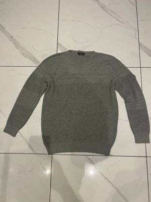 Grå Armani tröja - Snygg grå stickad tröja från Emporio Armani med rund hals och diskreta ribbade detaljer över bröstet och ärmarna. Patch med logga på vänster ärm ger en cool touch. Perfekt för dig som gillar stilrena och bekväma plagg.