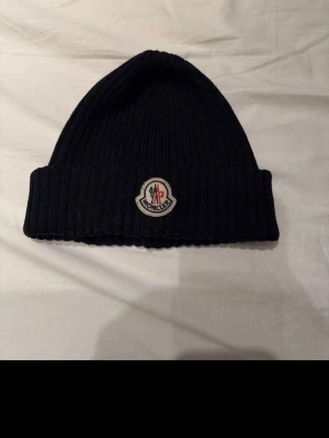 Mörkblå  ribbad Moncler mössa - Säljer en mörkblå ribbstickad mössa från Moncler med klassisk logotyp framtill. Mössan är tillverkad i mjukt material och har en uppvikt kant för extra stilpoäng. Perfekt för dig som vill ha en clean och snygg accessoar till vintern.