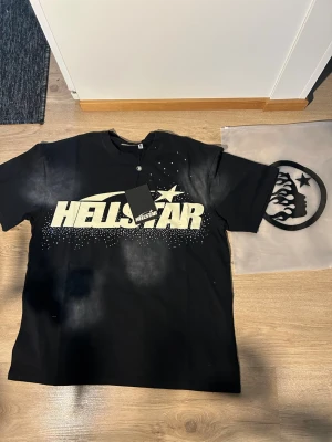 Svart Hellstar t-shirt med tryck - Svart t-shirt från Hellstar med stor vit logotyp och stjärnmotiv framtill, samt ett coolt ansikte med flames och stjärna på ryggen. T-shirten har rund hals och korta ärmar. Materialet är mjuk bomull och passformen är loose. Perfekt för dig som gillar streetwear och statement-plagg.