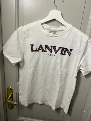 Vit Lanvin Paris t-shirt med tryck - Säljer en vit t-shirt från Lanvin Paris i storlek 14 passar xs. T-shirten har ett stort Lanvin-tryck på bröstet med färgglada detaljer i bokstäverna. Klassisk rund hals och korta ärmar. Materialet är mjuk bomull som känns skönt mot huden.