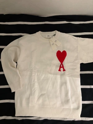 Vit stickad tröja från Ami med hjärta - Vit stickad tröja från Ami med ett stort rött hjärta och bokstaven A broderat på bröstet. Tröjan har rund hals, ribbade muddar och lång ärm. Perfekt för dig som gillar stilrena plagg med en twist. Snygg detalj framtill som sticker ut. Hör av dig om du undrar nått.