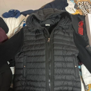 Moncler cardigan - Svart pufferjacka från Moncler med huva och ribbstickade ärmar. Jackan har quiltad front, två sidofickor med tryckknappar och dragkedja framtill. Moncler-logga på ärmen och snygga detaljer i både dun och stickad polyester. Perfekt för kalla dagar.