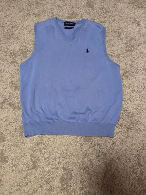 Blå stickad väst från Polo Ralph Lauren - Snygg ljusblå stickad väst från Polo Ralph Lauren med klassisk v-ringning och broderad logga på bröstet. Västen har ribbade kanter nedtill och runt halsen, perfekt att styla över en skjorta för en clean look.
