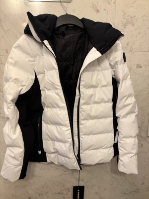 Vit/ svart pufferjacka för skidåkning Everest - Säljer en snygg pufferjacka från Everest  i vit och svart. Jackan har huva, dragkedja framtill och coola svarta detaljer längs sidorna och ärmarna, du kan även tajta till vid midjan i jackan som du ser på bilden. Märke syns på ärmen. Perfekt för skidåkning. Bär på 36 men tog 38 då man vill få plats med tjocka plagg under