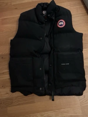 Svart dunväst från Canada Goose - Svart dunväst från Canada Goose med klassisk logotyp på bröstet. Västen har hög krage, två stora fickor framtill och dragkedja med tryckknappar. Perfekt för kalla dagar och har en stilren, tidlös look.