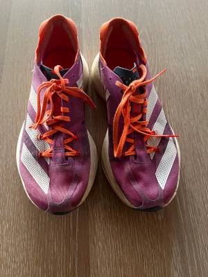 Adidas Adizero lAdios Pro löparskor - Säljer ett par Adidas Adizero löparskor i lila med vita ränder och orange detaljer. Skorna har platt sula, snörning och är tillverkade i lätt syntetmaterial med Lightstrike Pro-dämpning. Perfekta för dig som vill ha snygga och snabba sneakers till träningen.