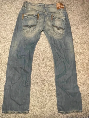 Replay jeans i ljusblå tvätt - Snygga Replay jeans i ljusblå tvätt med slitningar och coola detaljer på bakfickorna. Klassisk femficksmodell med raka ben och normal passform. Märkespatch bak i midjan och Replay-logga vid fickan. Perfekta för en avslappnad streetstil.