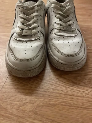 Nike Air Force 1 vita sneakers - Säljer ett par klassiska Nike Air Force 1 sneakers i helvitt skinn med perforerad tå och rund tå. Skorna har snörning och platt sula, samt den ikoniska Nike-loggan på sidan och under sulan. Perfekt för dig som gillar streetstyle och vill ha en tidlös look.