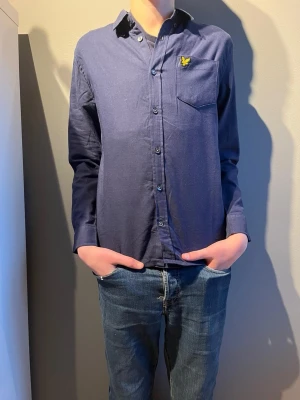 Mörkblå skjorta från Lyle & Scott - Snygg mörkblå skjorta från Lyle & Scott med klassisk krage och gul broderad logga på bröstfickan. Skjortan har knappar hela vägen och är långärmad. Materialet är mjukt och känns som bomull, perfekt för en chill och stilren look.