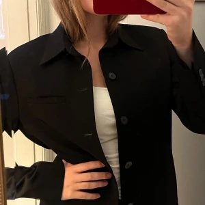 Svart vintagekavaj med knappar - Snygg svart kavaj med klassisk krage och raka linjer. Kavajen har knappar framtill och på ärmsluten, samt en diskret bröstficka. Passformen är normal och den är i väldigt fint material. Perfekt för en stilren look.