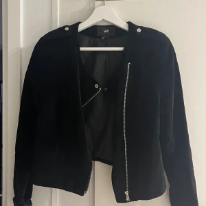 Svart mockajacka från H&M - Snygg svart mockajacka från H&M med silvriga dragkedjor och knappar på axlarna. Jackan har en klassisk bikerstil med sned dragkedja och lång ärm. Perfekt för dig som vill ha en stilren och cool look.