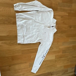 Vit half zip tröja Tommy Hilfiger - Beige långärmad tröja från Tommy Hilfiger med half zip och hög krage. Tröjan har logga broderad på bröstet och tryck längs ena ärmen. Tillverkad i mjuk bomull och har ribbade muddar vid ärmslut och krage.har endast legat i en garderob sedan den köptes nypris 899 kr.