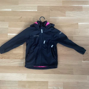 Svart vindjacka On The Peak - Svart vindjacka från On The Peak med huva och detaljer i rosa. Jackan har dragkedjor framtill och på fickor samt Aquatex Air Vent-tryck på ärmen. Perfekt för blåsiga dagar och har en sportig look.