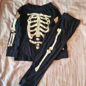 Tvådelad skelettdräkt som lyser i mörkret - Svart långärmad tröja och byxor med tryck av skelettben i vitt på framsidan och ärmarna. Perfekt för halloween eller temadagar. Tröjan är i mjuk bomull. Det är några ytterst små sprickor i trycket.