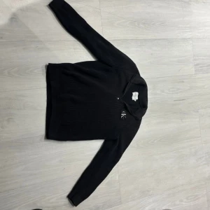 Svart stickad tröja Calvin Klein half zip - Svart stickad tröja från Calvin Klein med half zip och diskret CK-brodyr på bröstet. Tröjan har lång ärm, ribbade muddar och en högre krage. Perfekt för dig som gillar en clean och stilren look. Köpt den för 999kr har använt den 3- gånger 