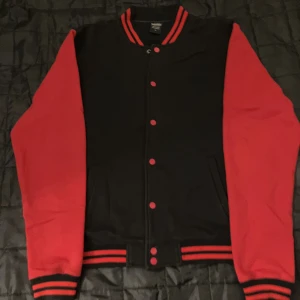 Svart och röd bomberjacka Urban Classics - Snygg svart och röd bomberjacka från Urban Classics med klassisk college-look. Jackan har röda ärmar, svarta muddar med röda ränder och knappar framtill. Perfekt för dig som vill sticka ut med en sportig och retro vibe.