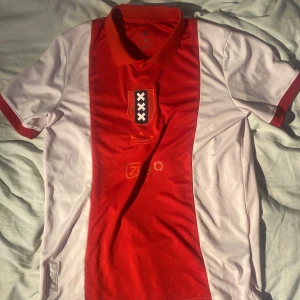 Ajax fotbollströja Adidas röd/vit S - Ajax fotbollströja från Adidas i klassisk röd och vit design med klubbens emblem och sponsortryck på bröstet. Tröjan har korta ärmar, pikékrage och är tillverkad i ett lätt och ventilerande material som passar perfekt för match eller träning.